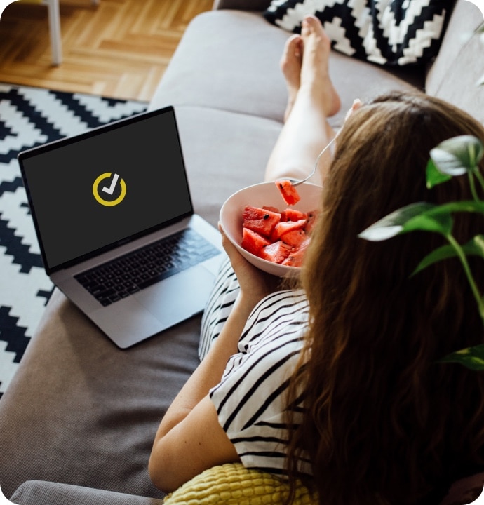 Kobieta jedząca arbuza na kanapie z laptopem, na którym jest uruchamiany program Norton 360 AntiVirus Plus